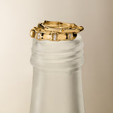 The Bamboo Ring in Gold sits elegantly on the frosted neck of a glass bottle, accented by a pair of gold hoop earrings against a neutral background for a minimalist and sophisticated display.