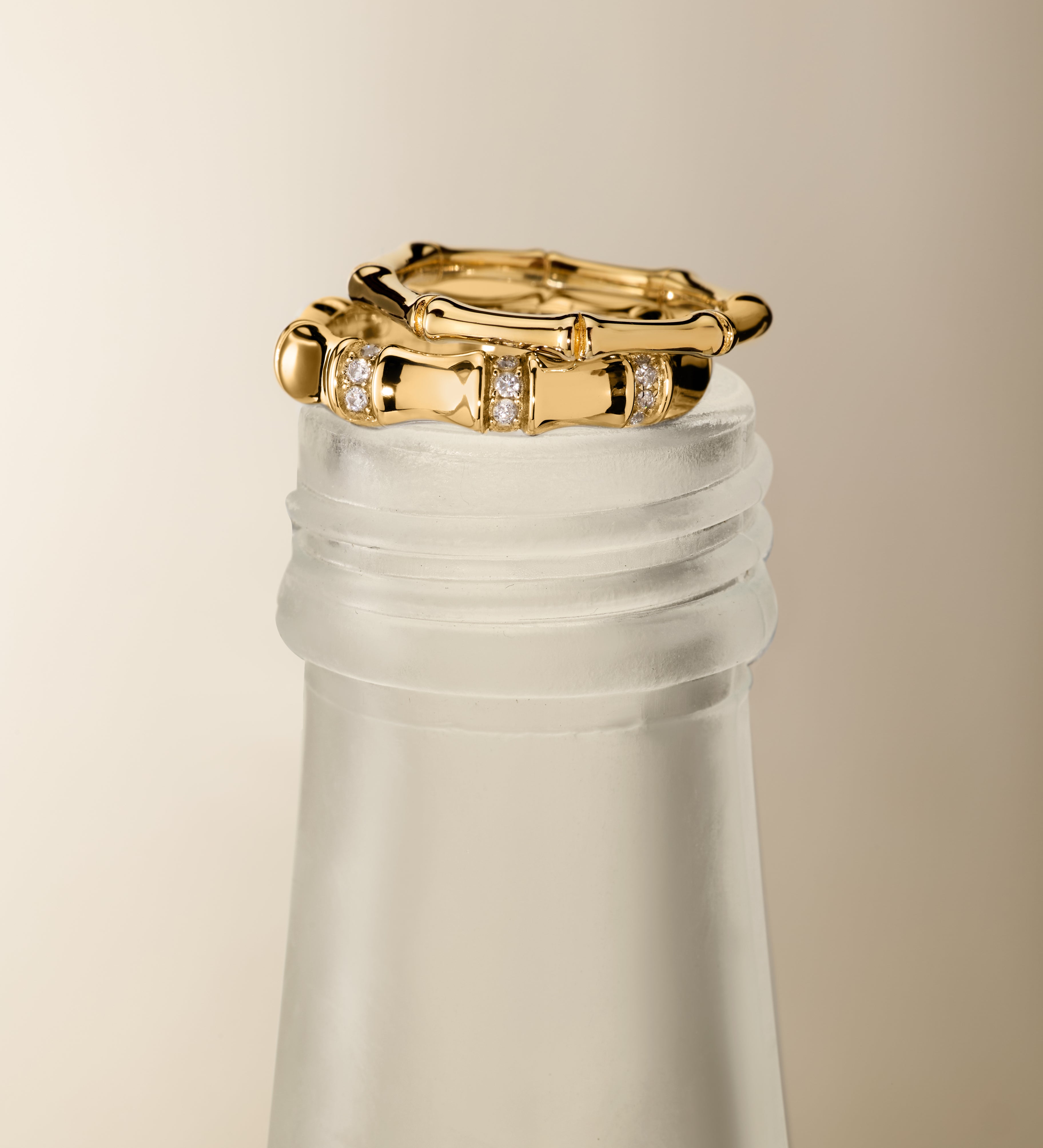 The Bamboo Ring in Gold sits elegantly on the frosted neck of a glass bottle, accented by a pair of gold hoop earrings against a neutral background for a minimalist and sophisticated display.