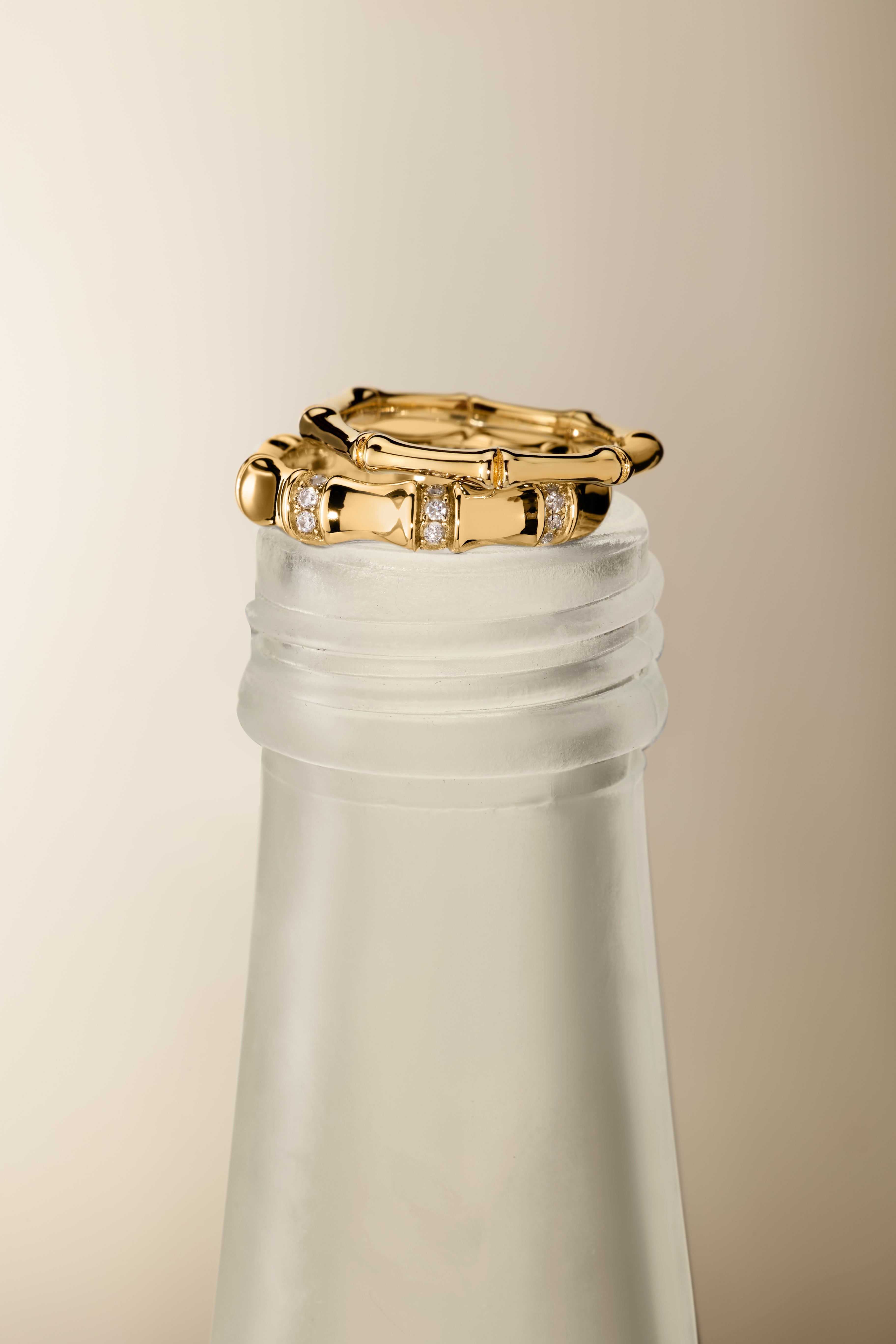The Bamboo Ring in Gold sits elegantly on the frosted neck of a glass bottle, accented by a pair of gold hoop earrings against a neutral background for a minimalist and sophisticated display.