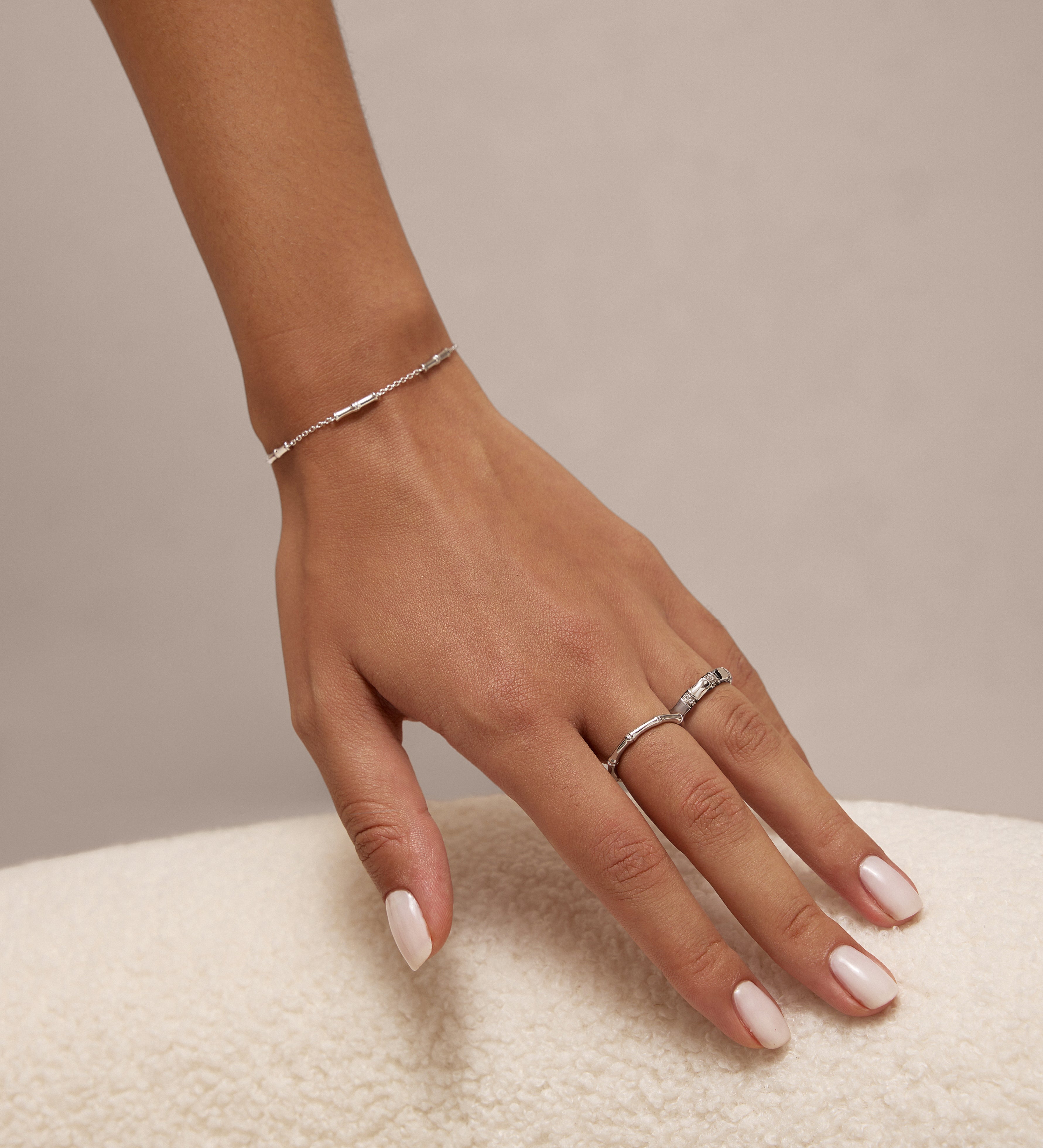 A hand with pale pink manicured nails rests on a soft, white textured surface, adorned with the Bamboo Chain Bracelet in Silver and two S925 sterling silver rings on the middle and ring fingers.