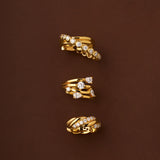 The Vina Crystal Bezel Ear Cuff in Gold, featuring sparkling cubic zirconia and both smooth and textured gold finishes, is elegantly displayed on a brown background.