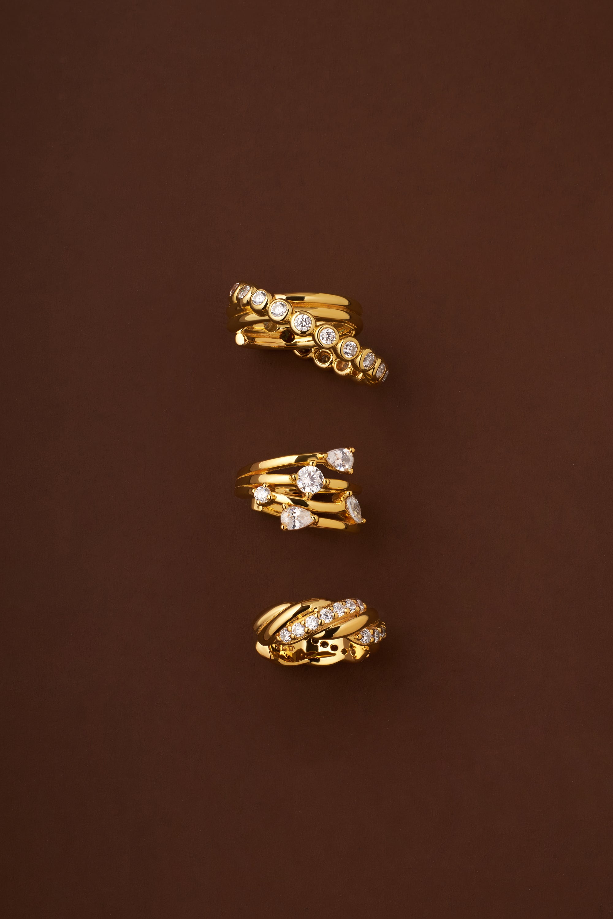 The Vina Crystal Bezel Ear Cuff in Gold, featuring sparkling cubic zirconia and both smooth and textured gold finishes, is elegantly displayed on a brown background.
