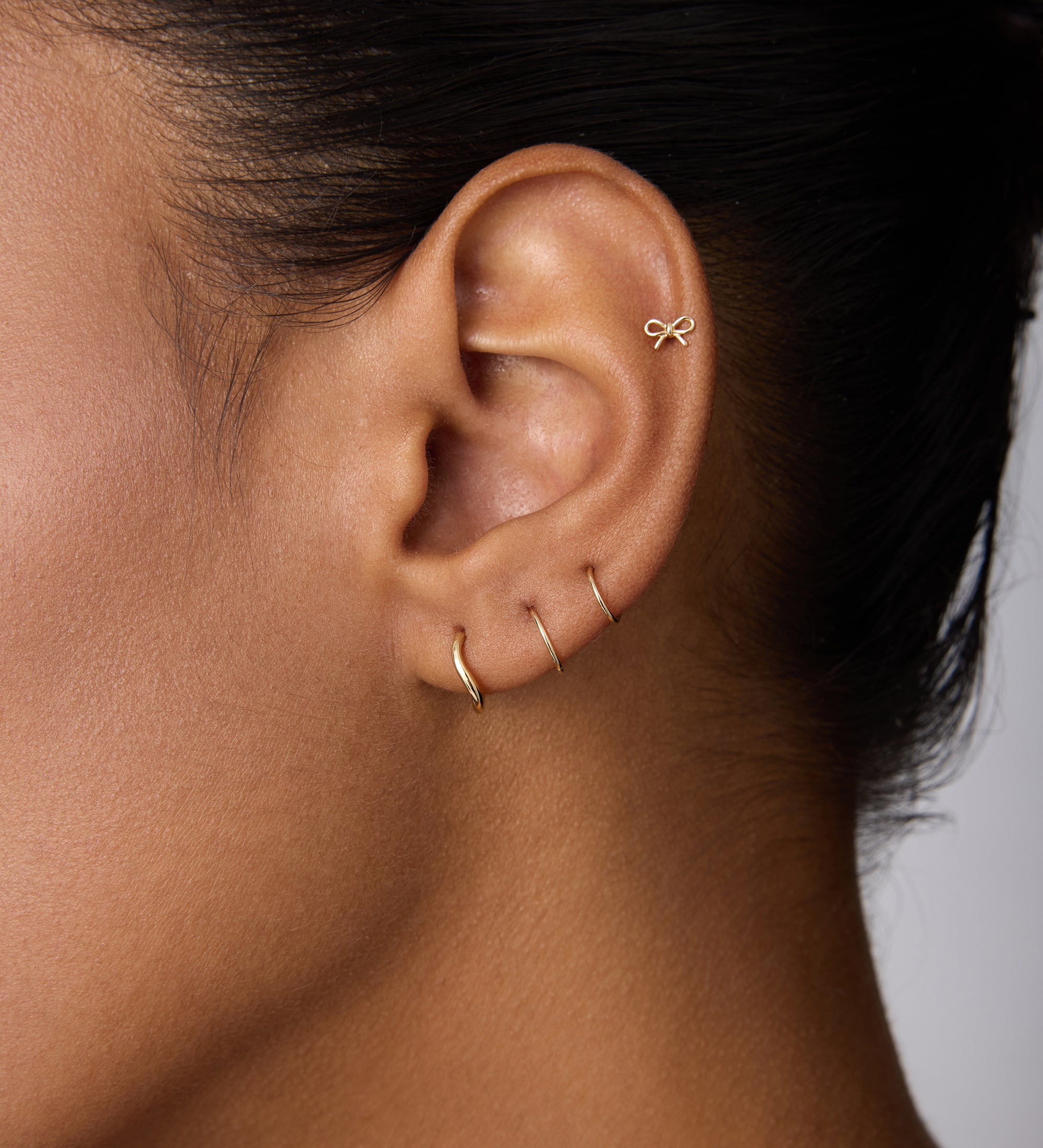 Close-up of an ear adorned with four gold earrings: a 14K Solid Yellow Gold Bow Tie Flat Back Stud at the top and three minimalist hoops along the earlobe. The person has smooth skin and dark hair pulled back.