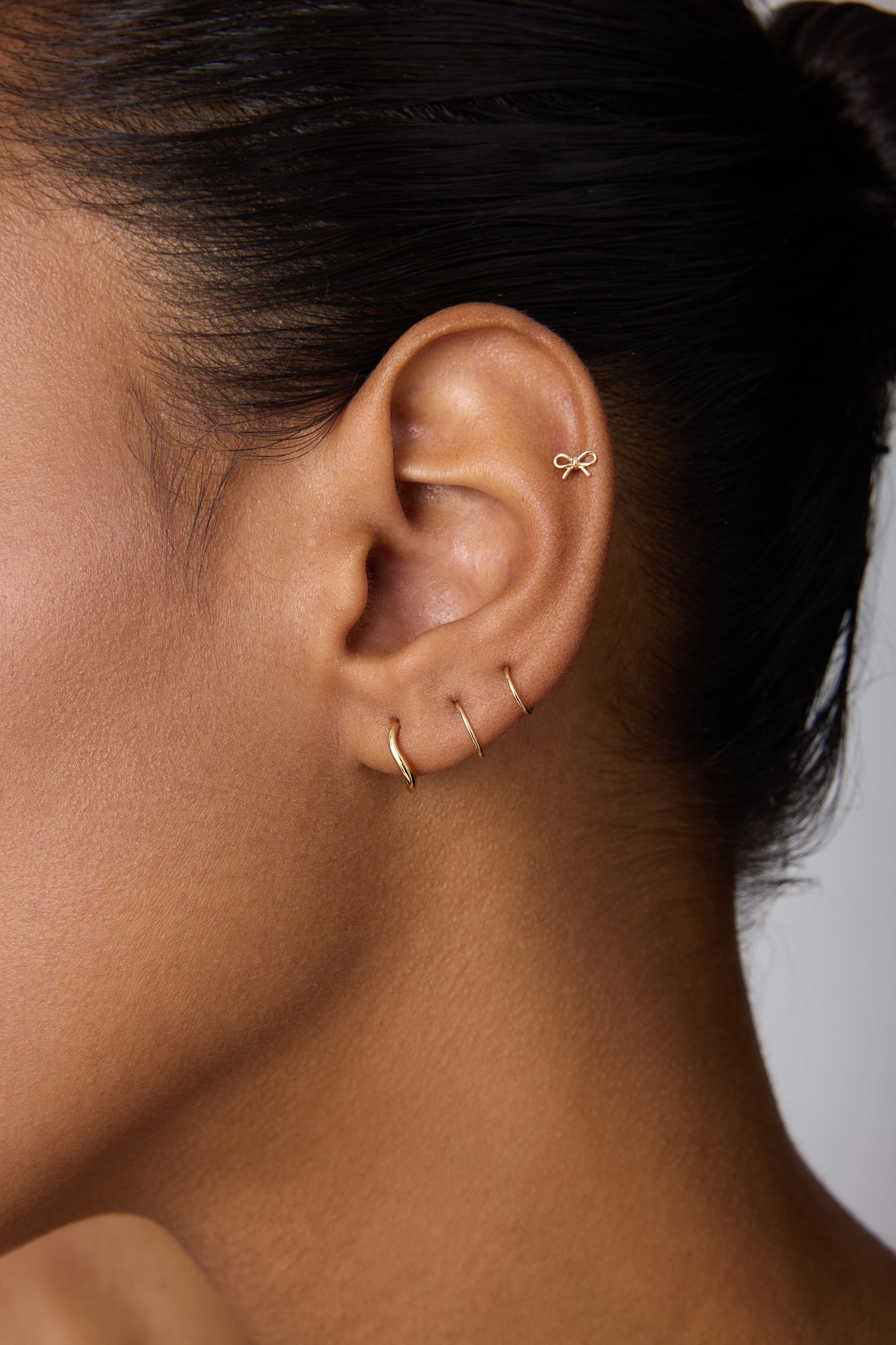 Close-up of an ear adorned with four gold earrings: a 14K Solid Yellow Gold Bow Tie Flat Back Stud at the top and three minimalist hoops along the earlobe. The person has smooth skin and dark hair pulled back.
