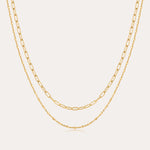The Mini Paperclip Double Chain Necklace in Gold features two 18k gold plated layered chains—one with delicate elongated links, the other with fine round links—displayed elegantly against a plain, light background.