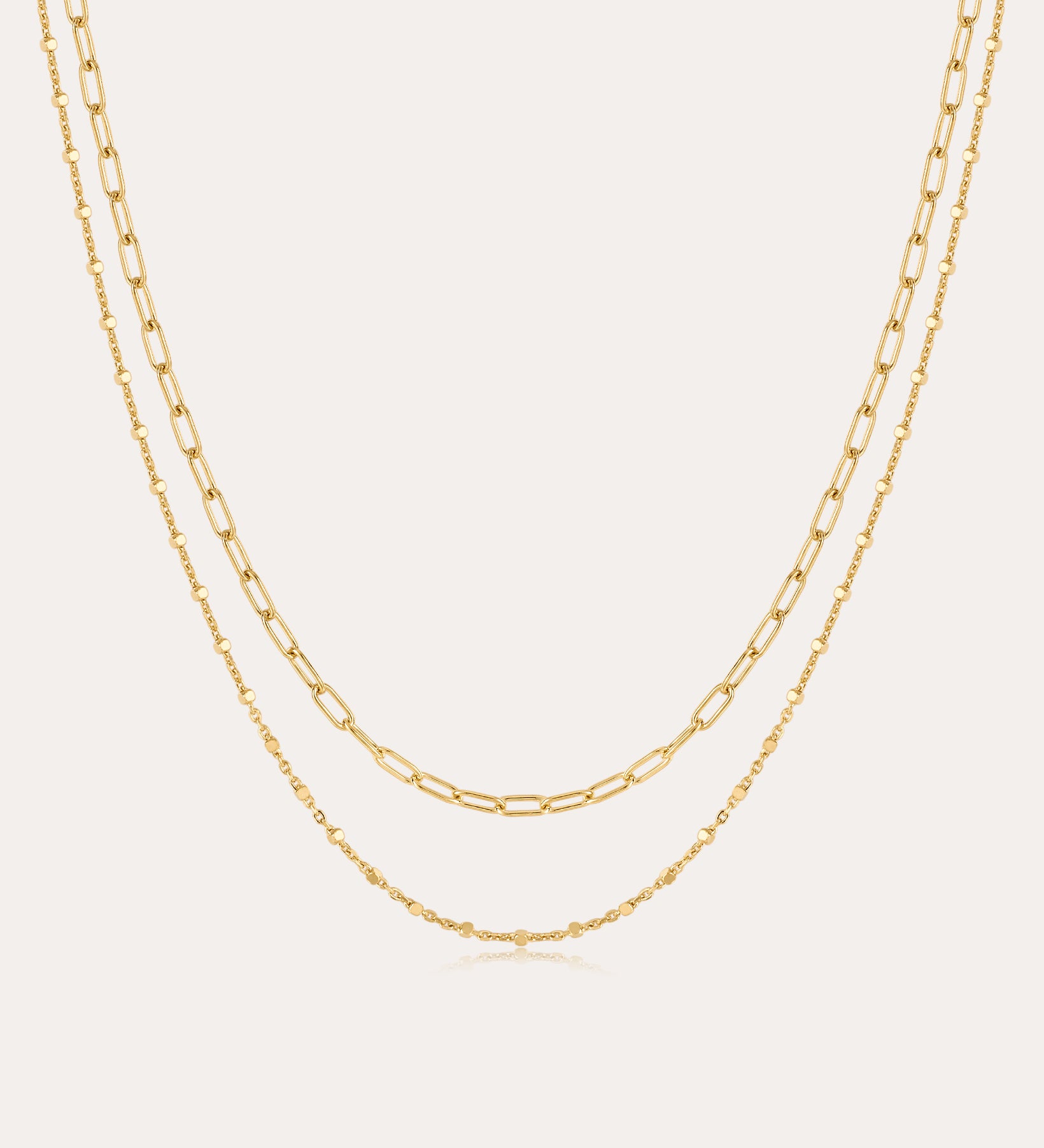 The Mini Paperclip Double Chain Necklace in Gold features two 18k gold plated layered chains—one with delicate elongated links, the other with fine round links—displayed elegantly against a plain, light background.