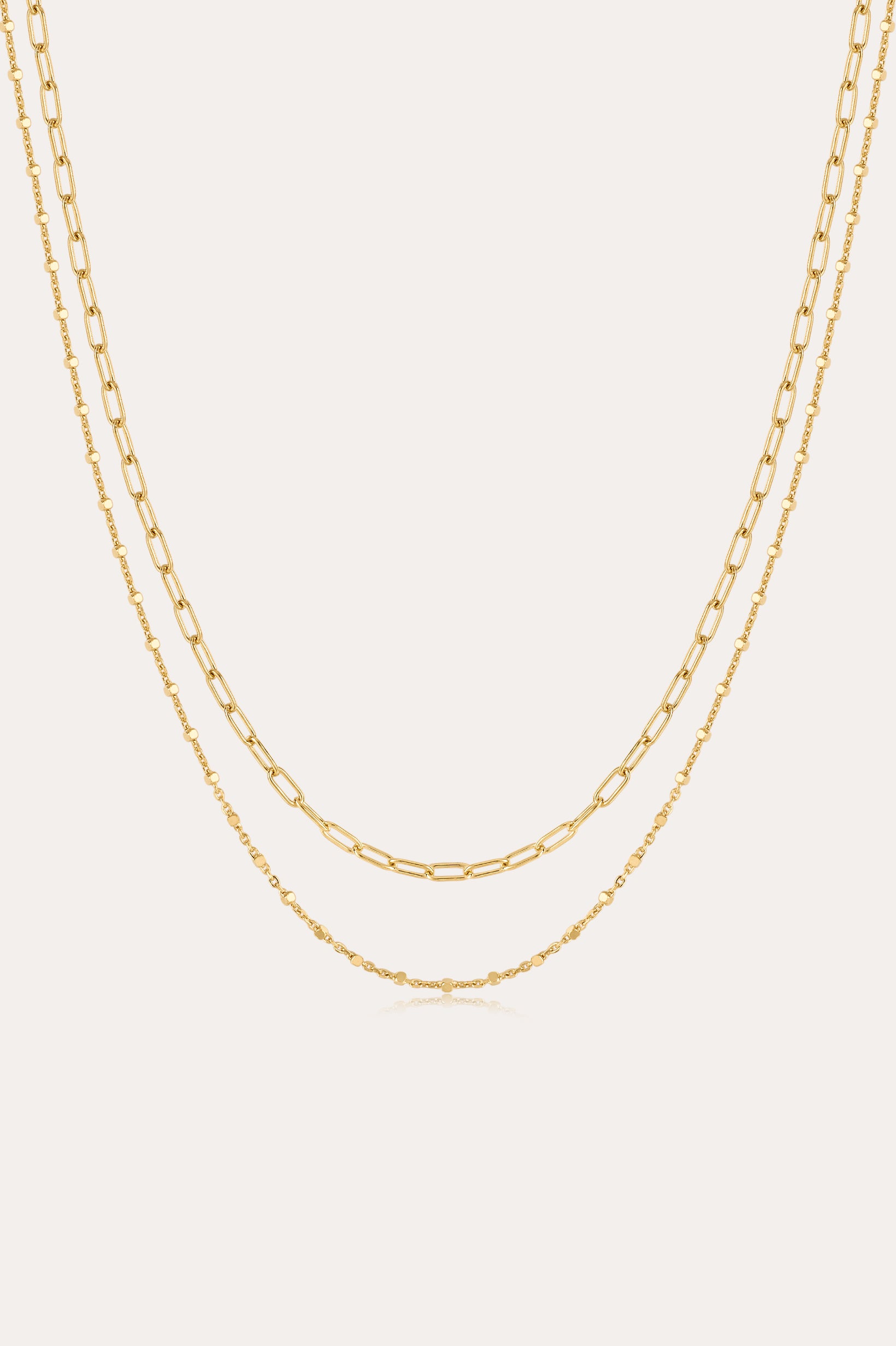 The Mini Paperclip Double Chain Necklace in Gold features two 18k gold plated layered chains—one with delicate elongated links, the other with fine round links—displayed elegantly against a plain, light background.