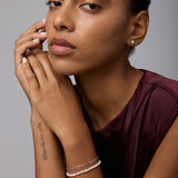 A woman with a neutral expression poses against a plain background. She wears a sleeveless maroon top, bracelets, and Freshwater Pearl Studs in Silver. A tattoo marks her forearm as her hands gently touch her face and chin.