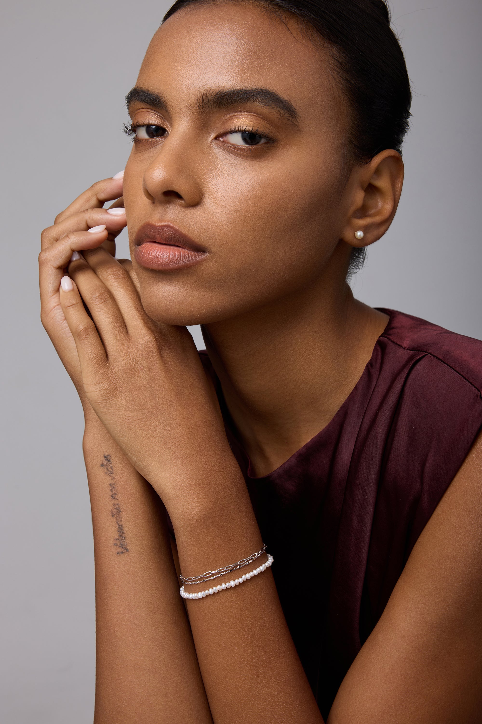 A woman with a neutral expression poses against a plain background. She wears a sleeveless maroon top, bracelets, and Freshwater Pearl Studs in Silver. A tattoo marks her forearm as her hands gently touch her face and chin.