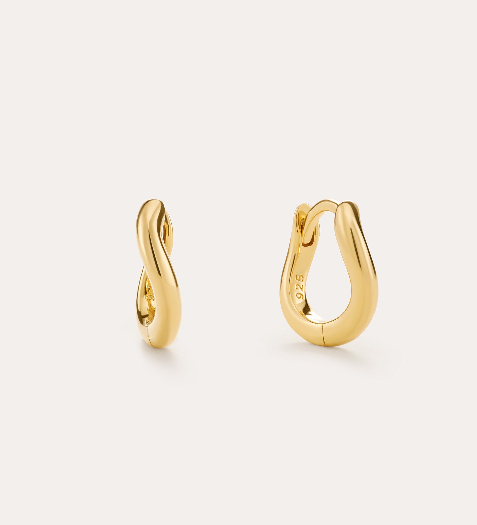 The Nori Mini Twisted Huggies in Gold are 18k gold plated earrings with a smooth, twisted design, beautifully shown on a light beige background.
