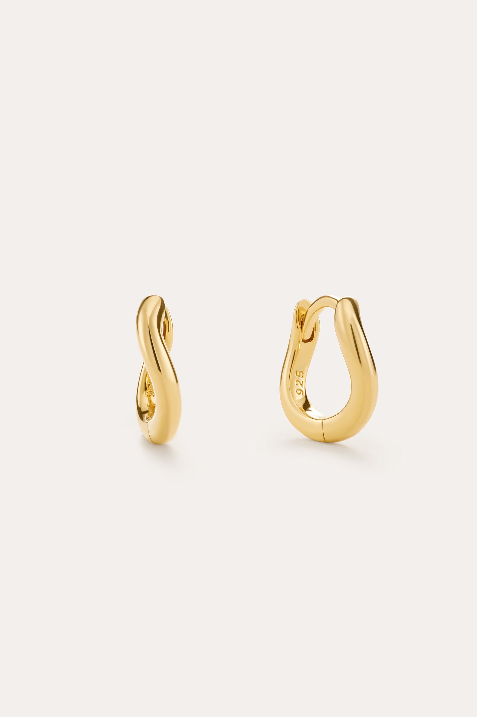 The Nori Mini Twisted Huggies in Gold are 18k gold plated earrings with a smooth, twisted design, beautifully shown on a light beige background.