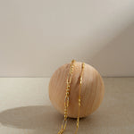 The Carly Paperclip Chain Bead Bracelet in Gold rests gracefully on a smooth, round wooden sphere, highlighted by a neutral beige background with soft lighting and gentle shadows.