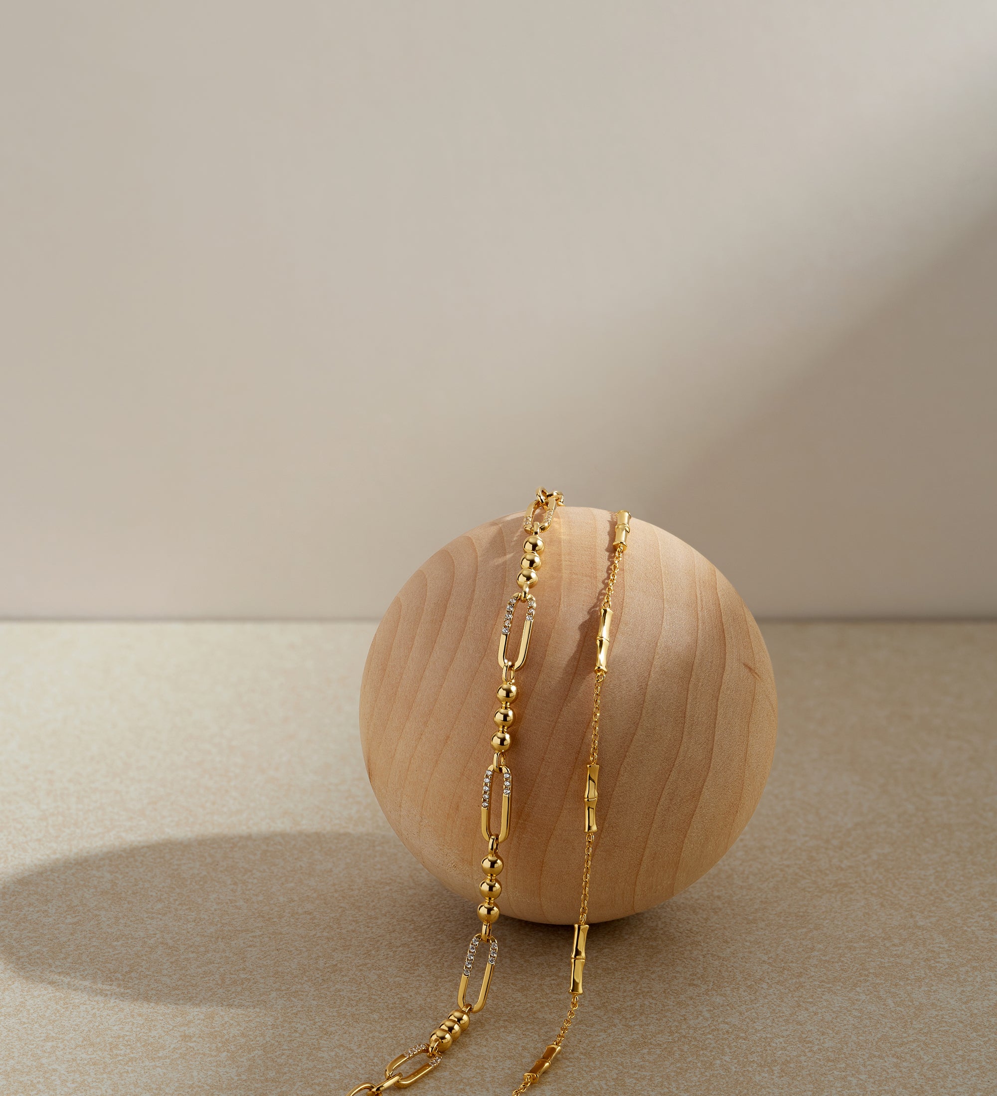 The Carly Paperclip Chain Bead Bracelet in Gold rests gracefully on a smooth, round wooden sphere, highlighted by a neutral beige background with soft lighting and gentle shadows.