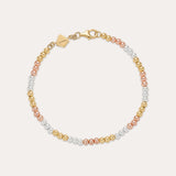 The Cecelia Three Tone Trinity Bracelet features small gold, sterling silver, and rose gold beads in a gradient pattern with a gold clasp and tag. This dainty bracelet is 18k gold plated and styled on a light background.