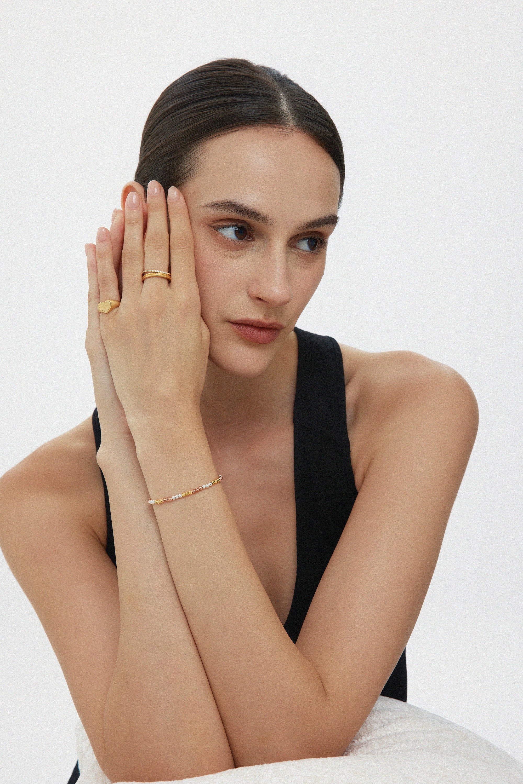A woman with brown hair in a low bun, wearing a black sleeveless top, rests her chin on folded hands. She wears gold and yellow gemstone rings and the Cecelia Three Tone Trinity Bracelet with colored stones.