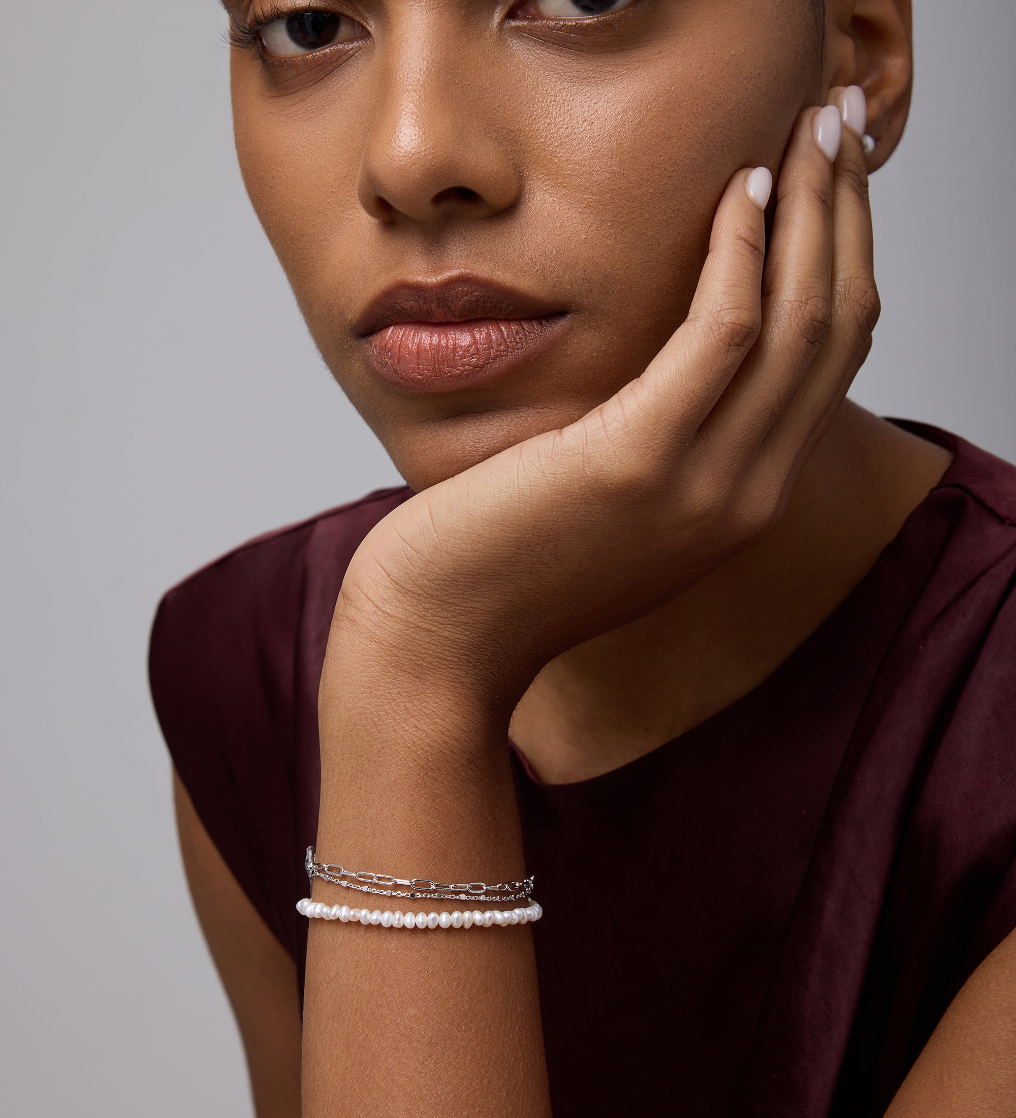 A woman with medium skin tone rests her chin on her hand, wearing a sleeveless burgundy top and the Celia Micro Freshwater Pearl Bracelet in Silver. She gazes thoughtfully at the camera with a neutral expression.