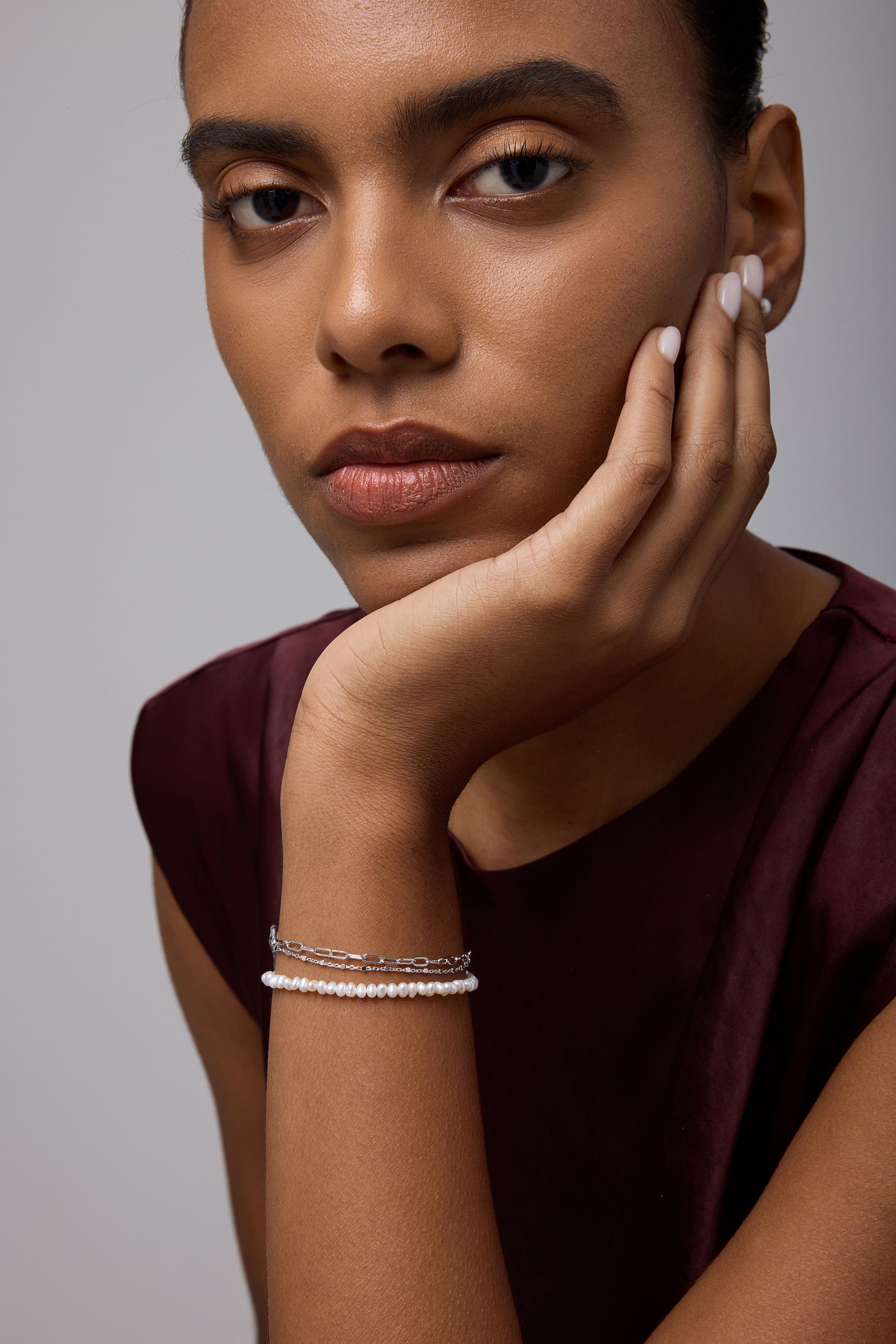 A woman with medium skin tone rests her chin on her hand, wearing a sleeveless burgundy top and the Celia Micro Freshwater Pearl Bracelet in Silver. She gazes thoughtfully at the camera with a neutral expression.