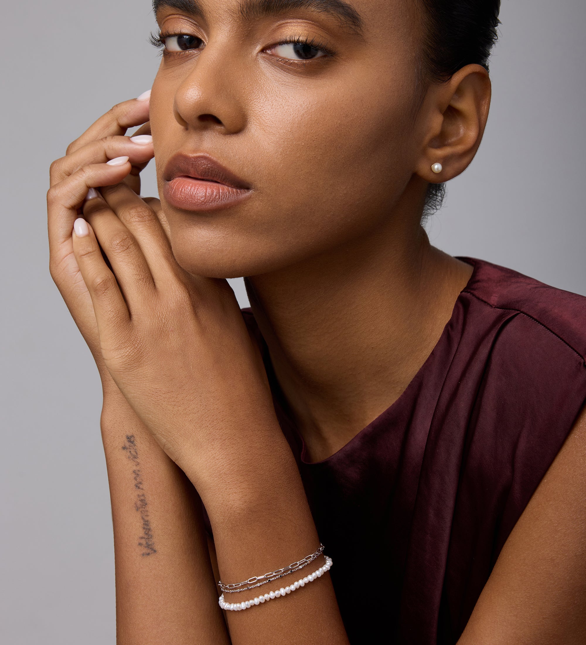 A woman with smooth, medium-brown skin wears a sleeveless burgundy top and stud earrings. She shows off her forearm tattoo and the Mini Paperclip Double Chain Bracelet in Silver, hands near her face against a plain background.