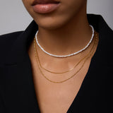 A close-up of a woman wearing a black blazer and layered necklaces: the Celia Micro Freshwater Pearl Choker Necklace in Gold and two thin gold chains, with only her lower face, neck, and part of her chest visible.