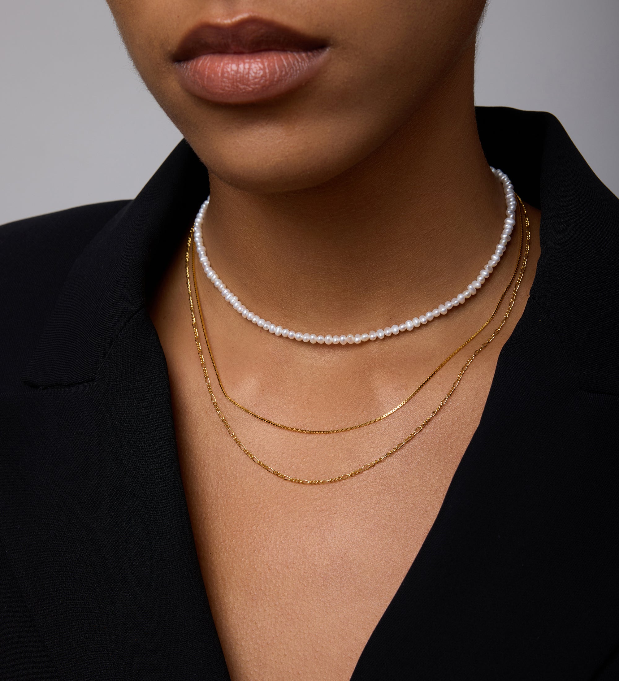 A close-up of a woman wearing a black blazer and layered necklaces: the Celia Micro Freshwater Pearl Choker Necklace in Gold and two thin gold chains, with only her lower face, neck, and part of her chest visible.