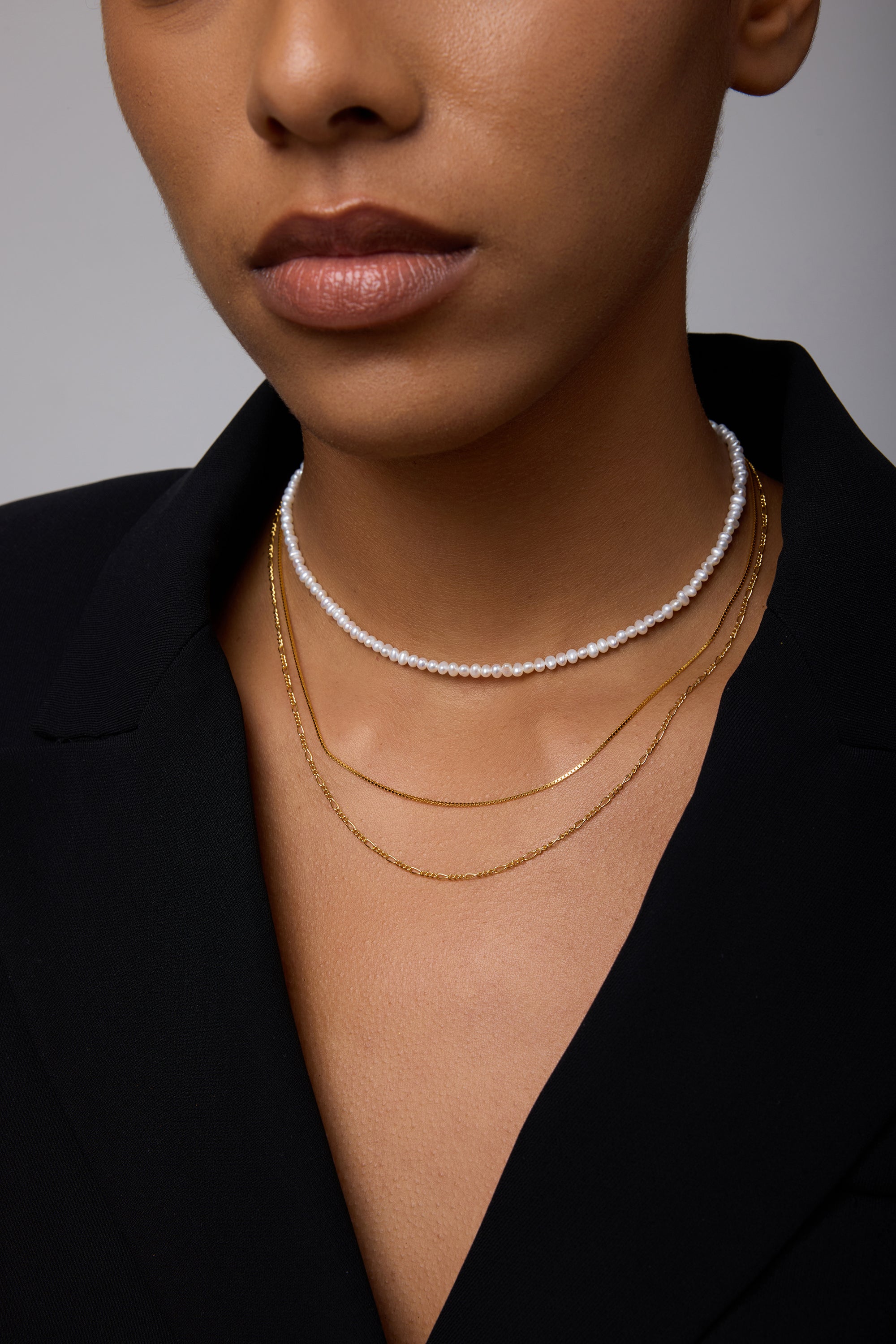 A close-up of a woman wearing a black blazer and layered necklaces: the Celia Micro Freshwater Pearl Choker Necklace in Gold and two thin gold chains, with only her lower face, neck, and part of her chest visible.