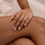 A woman with light pink nails and a gold ring rests her hand on her crossed leg, also wearing the Charlotte Eternity Ridged Ring in Gold. She is dressed in a sleeveless, light plaid shirt with white buttons.