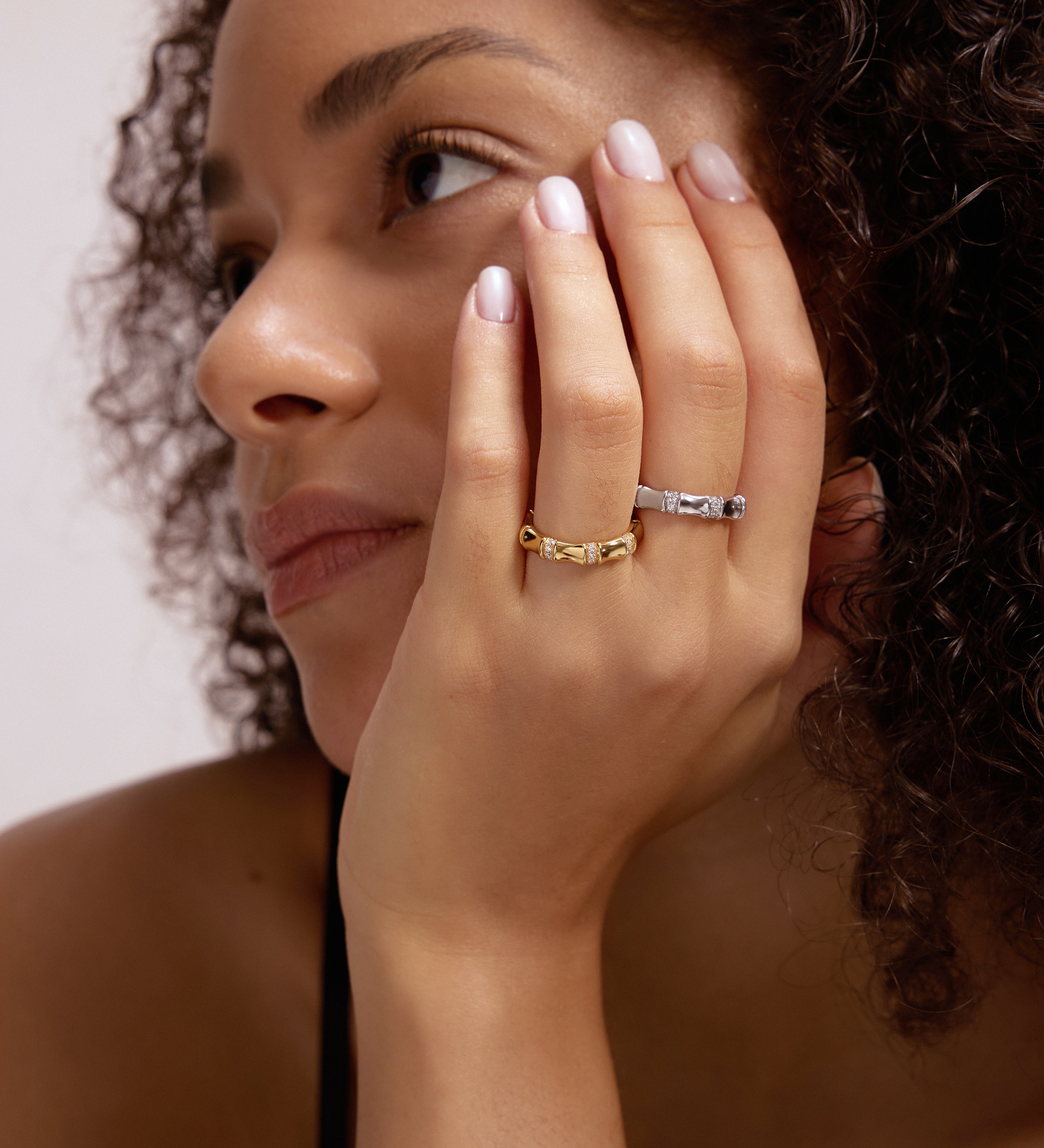 A woman with curly hair, natural makeup, and a thoughtful expression rests her chin on her hand, showcasing the Bamboo Chunky Crystal Ring in Gold.