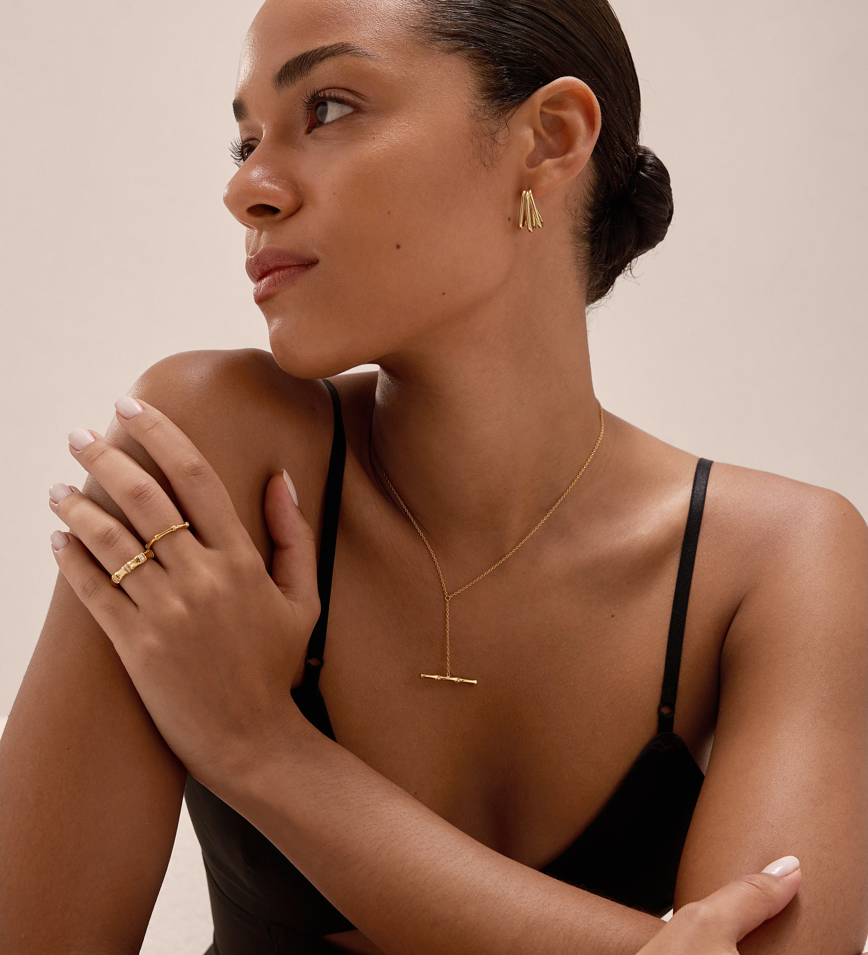 A woman with smooth, pulled-back hair wears a black camisole and gold jewelry, including earrings, a necklace, bracelet, and the Bamboo Chunky Crystal Ring in Gold. She looks to the side with one arm crossed over her chest.