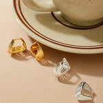 A beige cup and saucer with coffee sit on a light surface. In front, two pairs of modern earrings are displayed, including the Juno Ridged Chunky Hoops in Gold and a pair of silver geometric earrings.