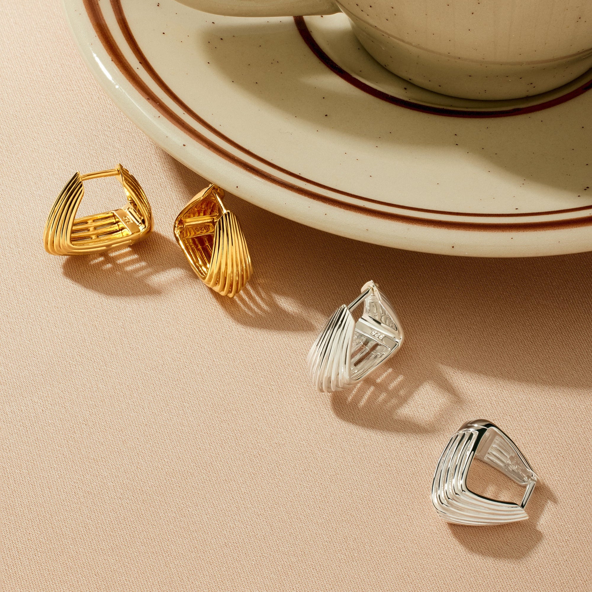 A beige cup and saucer with coffee sit on a light surface. In front, two pairs of modern earrings are displayed, including the Juno Ridged Chunky Hoops in Gold and a pair of silver geometric earrings.