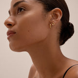 A woman with smooth skin and dark hair in a low bun wears the Colette Droop Link Huggie Earrings in Gold and a black top, gazing upward against a neutral background.