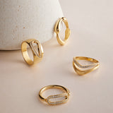 Four gold rings, including the striking Colette Link Droop Ring in Gold with its geometric design and small diamonds, are displayed on a pale surface, with one ring leaning against a textured, cone-shaped object.