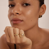 A woman with smooth, glowing skin and natural makeup rests her chin on her hand, showing off gold rings—including the Colette Link Droop Ring in Gold—and a small gold hoop earring. She gazes calmly at the camera against a neutral background.