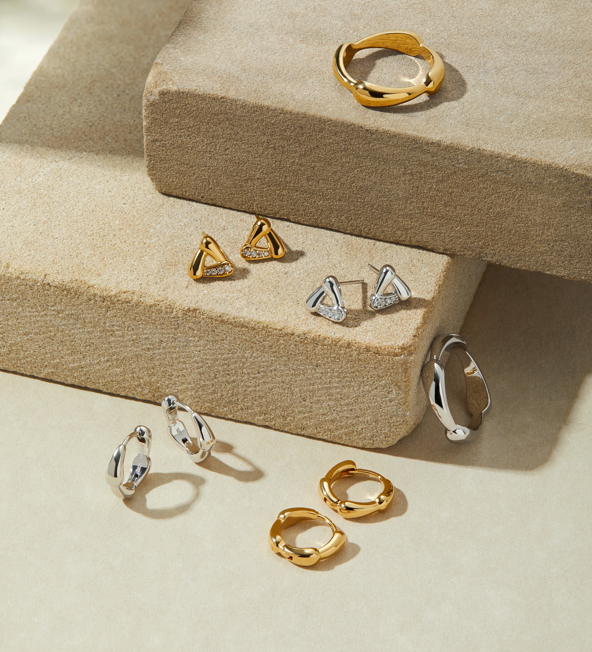 A display of gold and silver jewelry, featuring rings, hoop earrings, and the Colette Link Droop Ring in Gold, arranged on textured beige platforms under soft lighting.