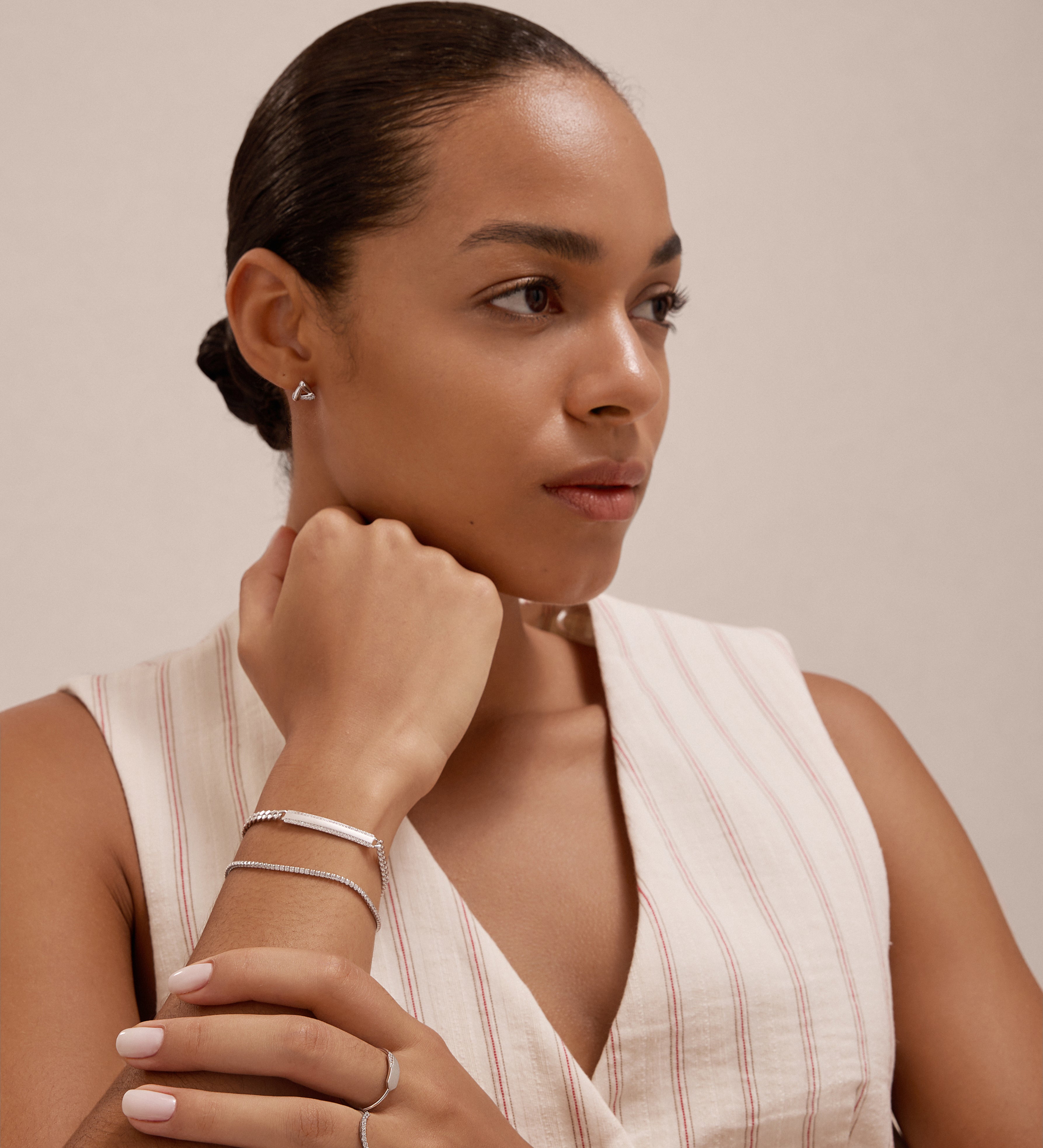 A woman with neatly styled hair, wearing a sleeveless pinstriped top and minimalist rings and bracelets, poses thoughtfully. She accessorizes with the Colette Droop Link Studs in Silver for a subtle, modern accent.