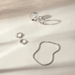 Four silver rings, including the Colette Link Droop Ring in Silver, and a silver bracelet are arranged on a cream-colored surface with soft, natural shadows—an elegant display of skate stopper jewelry.