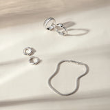 Four silver rings, including the Colette Link Droop Ring in Silver, and a silver bracelet are arranged on a cream-colored surface with soft, natural shadows—an elegant display of skate stopper jewelry.