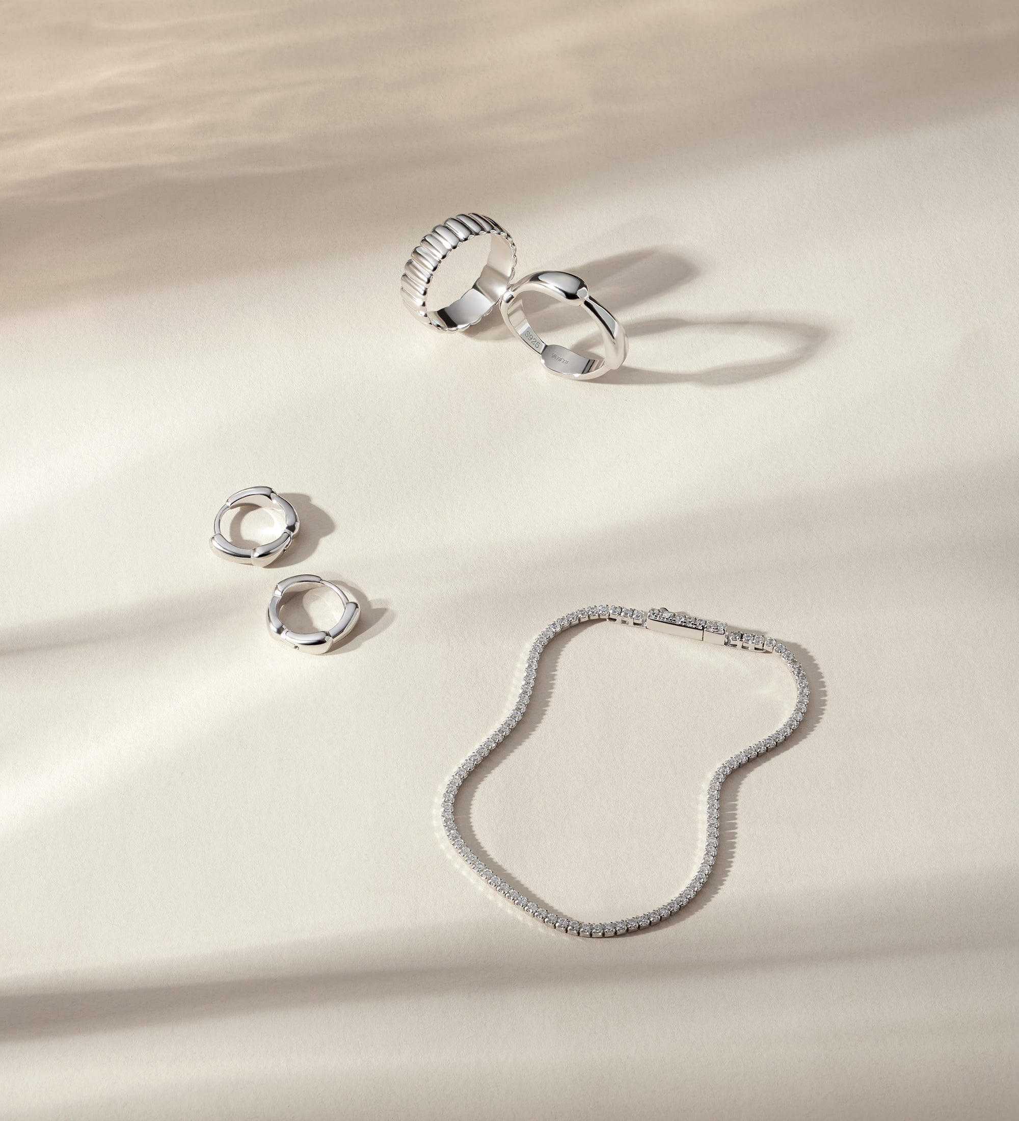 Four silver rings, including the Colette Link Droop Ring in Silver, and a silver bracelet are arranged on a cream-colored surface with soft, natural shadows—an elegant display of skate stopper jewelry.