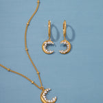 The Crescent Moon Crystal Charm Huggies in Gold, featuring crescent pendants adorned with sparkling cubic zirconia stones, are displayed on a light blue background.