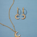 The Crescent Moon Crystal Charm Huggies in Gold, featuring crescent pendants adorned with sparkling cubic zirconia stones, are displayed on a light blue background.