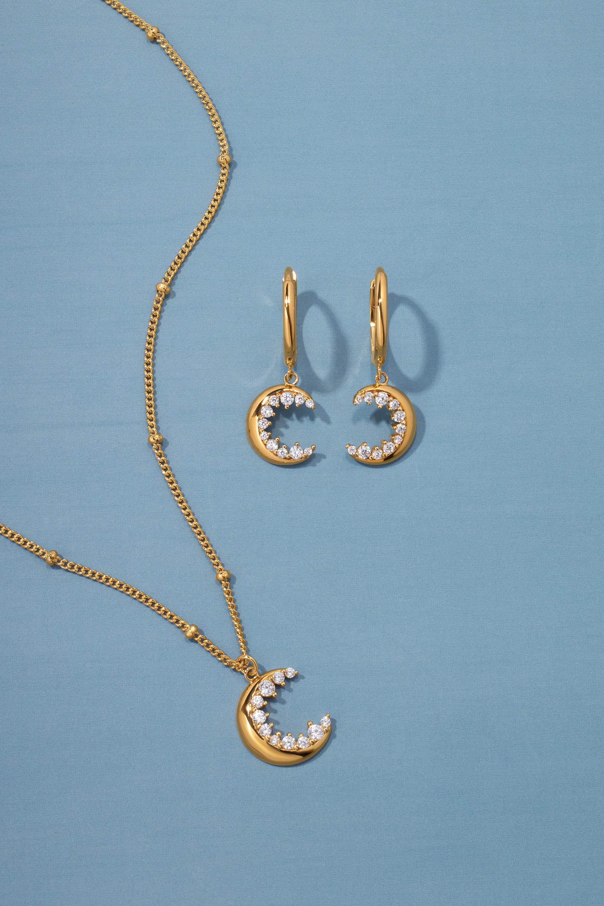 The Crescent Moon Crystal Charm Huggies in Gold, featuring crescent pendants adorned with sparkling cubic zirconia stones, are displayed on a light blue background.