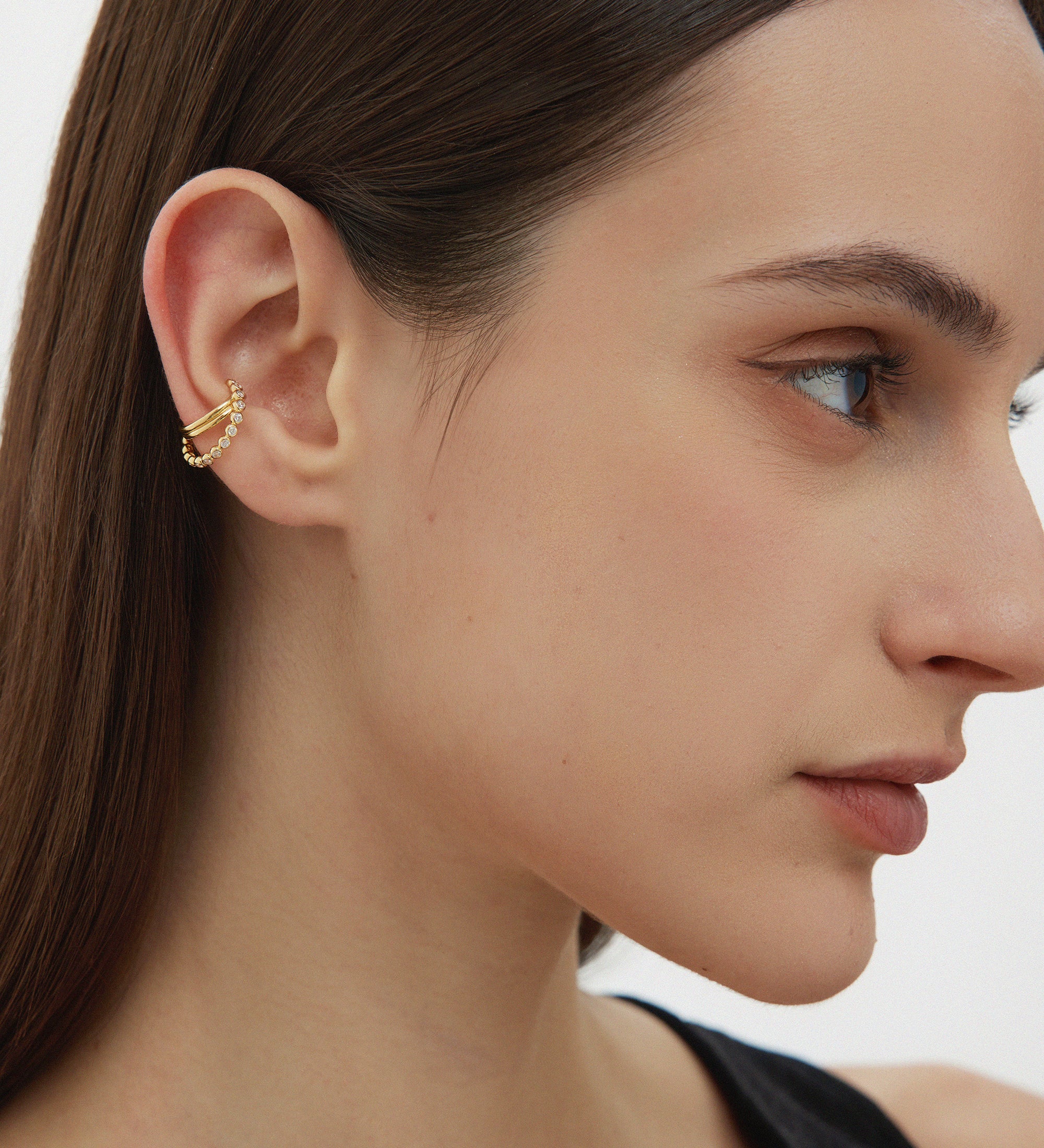 A woman with straight brown hair wears the Vina Crystal Bezel Ear Cuff in Gold, featuring cubic zirconia on her upper ear. She sports natural makeup and a black top, set against a plain background.