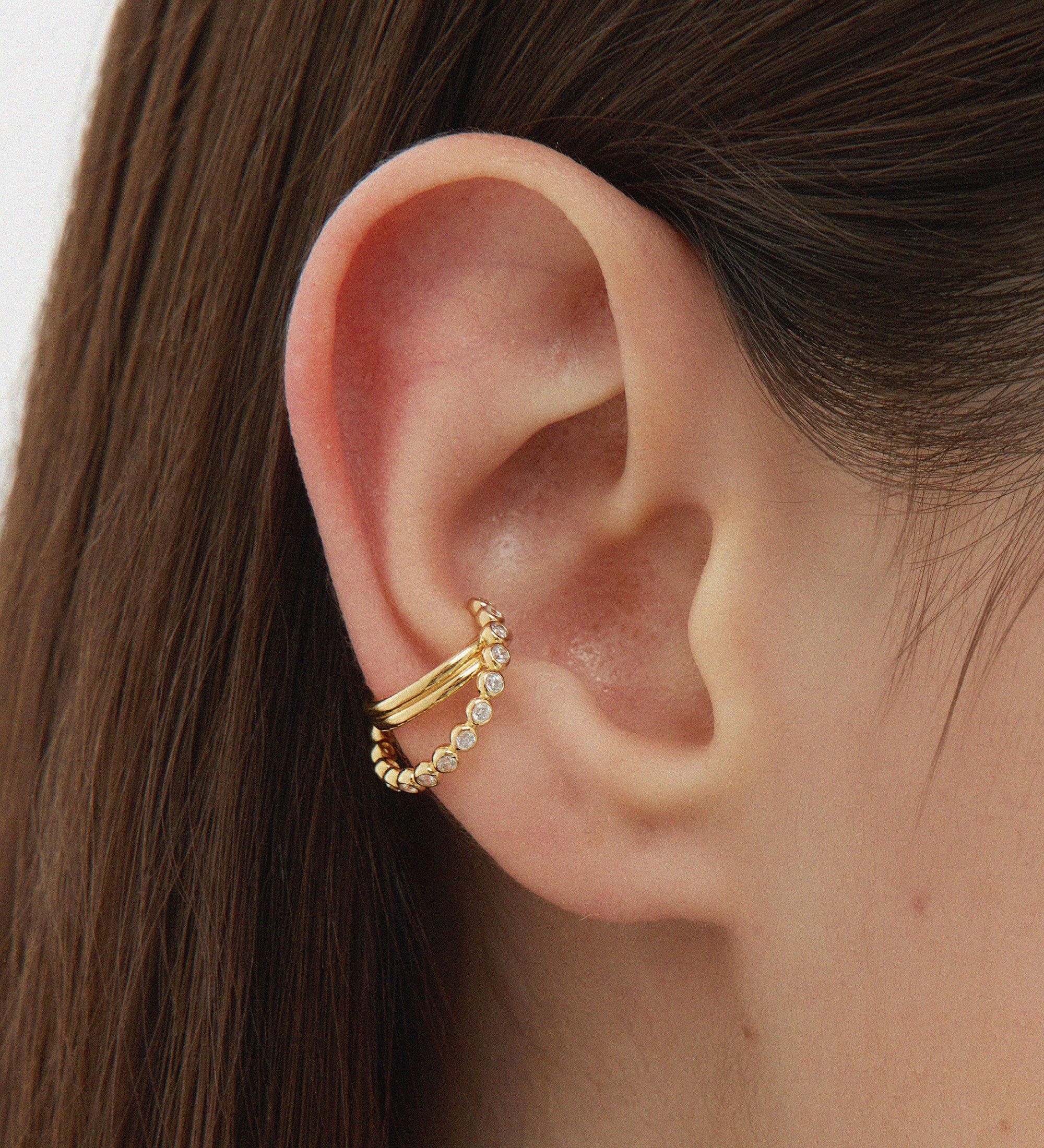 Close-up of a person’s ear with two gold ear cuffs, one smooth and one beaded. The person has straight brown hair and fair skin. Add sparkle by pairing these cuffs with the Vina Crystal Bezel Ear Cuff in Gold.