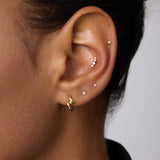 A close-up of a person’s ear with medium brown skin and dark hair wearing multiple piercings, including the 14K Solid White Gold Lab-grown Diamond Prong Set Flat Back Stud - 1.5mm in the lobe alongside gold and silver studs.
