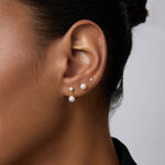 Close-up of a person’s ear wearing three earrings: a 14K solid yellow gold lab-grown diamond prong set flat back stud (2mm), a medium pearl stud, and a gold hoop with dangling pearl. The person has smooth skin and dark hair pulled back.