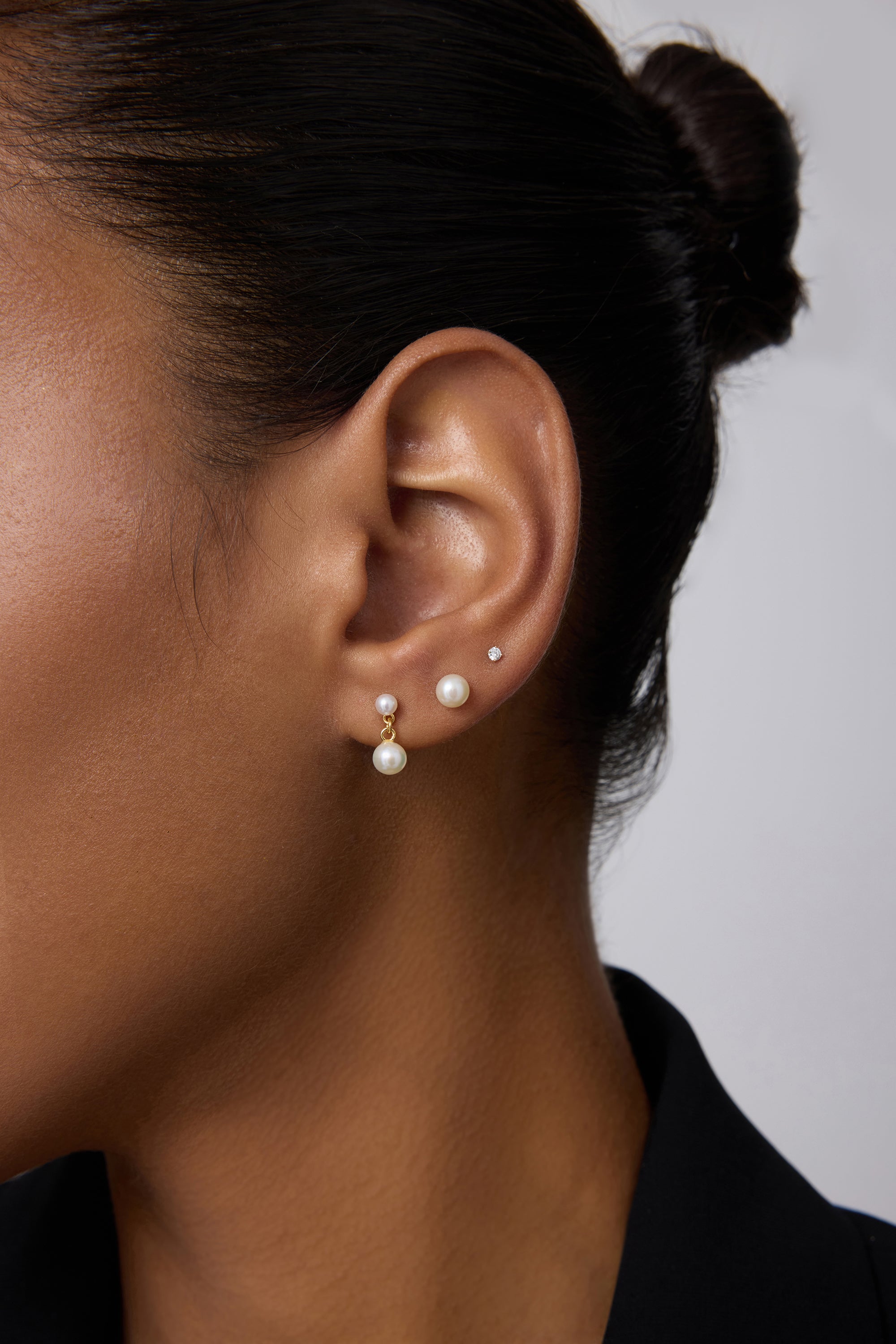 Close-up of a person’s ear wearing three earrings: a 14K solid yellow gold lab-grown diamond prong set flat back stud (2mm), a medium pearl stud, and a gold hoop with dangling pearl. The person has smooth skin and dark hair pulled back.