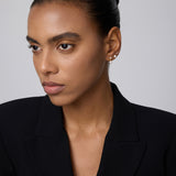A woman with smooth brown skin and dark hair pulled back wears a black blazer and a 14K solid yellow gold lab-grown diamond prong set flat back stud (2mm), looking slightly to the side against a plain light background.