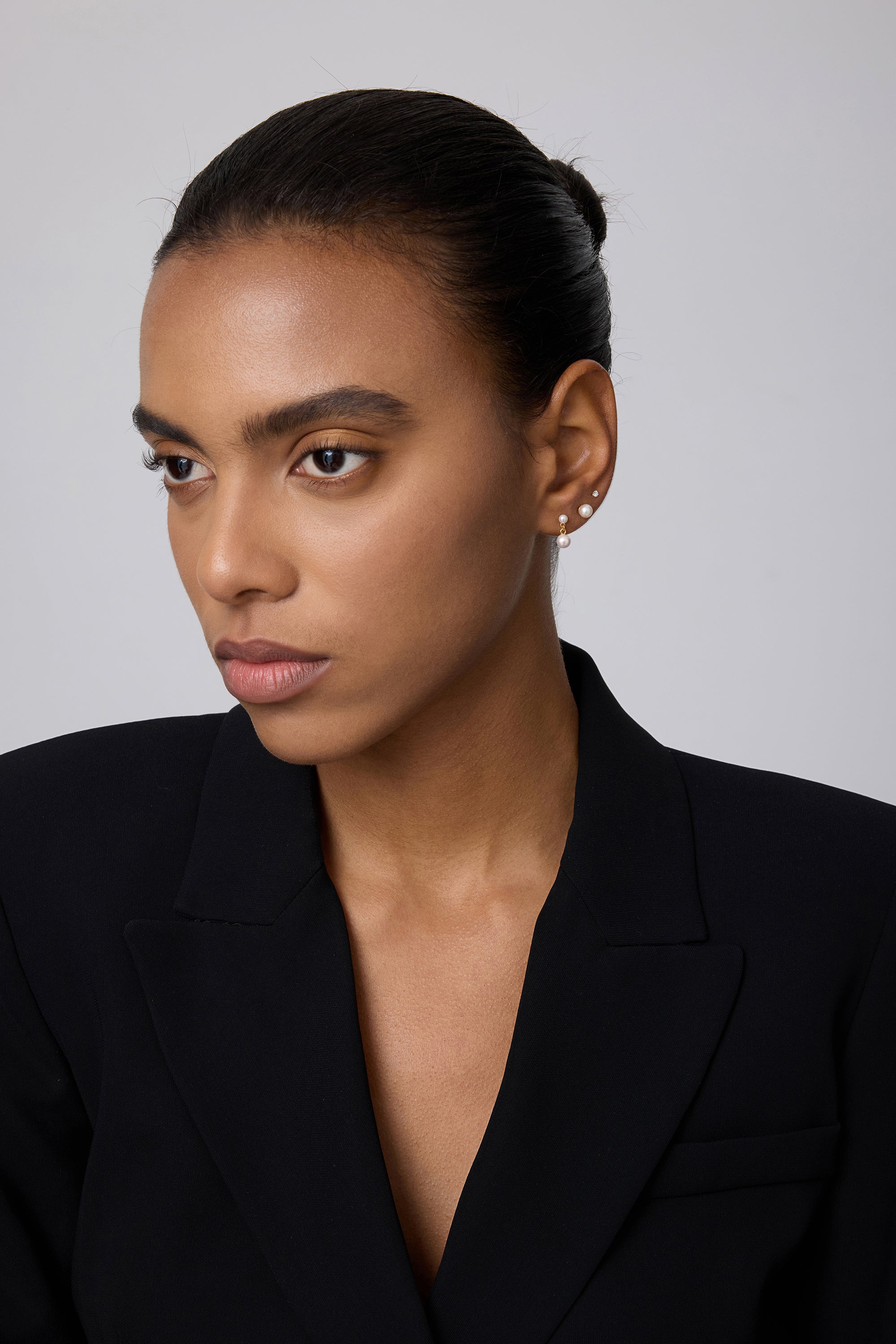 A woman with smooth brown skin and dark hair pulled back wears a black blazer and a 14K solid yellow gold lab-grown diamond prong set flat back stud (2mm), looking slightly to the side against a plain light background.
