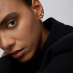 A close-up of a person’s face in a black outfit, highlighting smooth skin, groomed brows, neutral makeup, and three earrings—one is the 14K Solid Yellow Gold Lab-grown Diamond Prong Set Flat Back Stud (2mm)—against a plain light background.