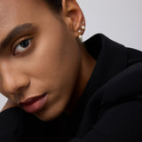 A close-up of a person’s face in a black outfit, highlighting smooth skin, groomed brows, neutral makeup, and three earrings—one is the 14K Solid Yellow Gold Lab-grown Diamond Prong Set Flat Back Stud (2mm)—against a plain light background.